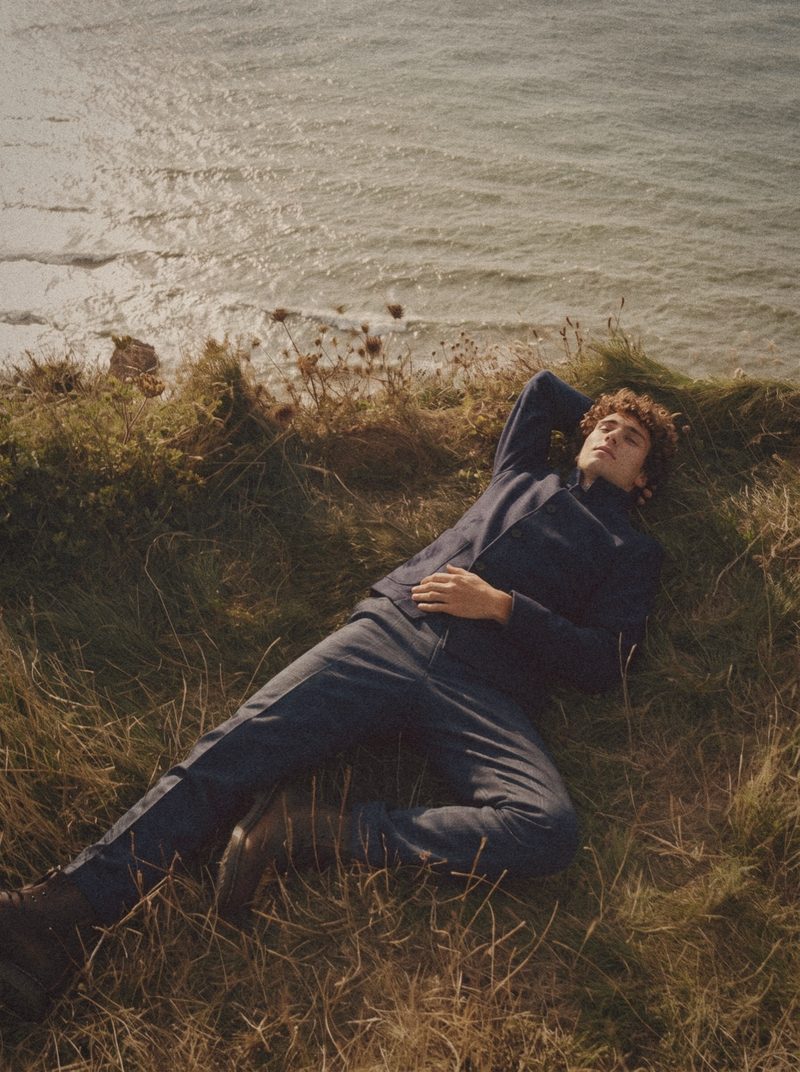 A man in a navy shirt jacket lies in the grass at a cliff edge above the sea.