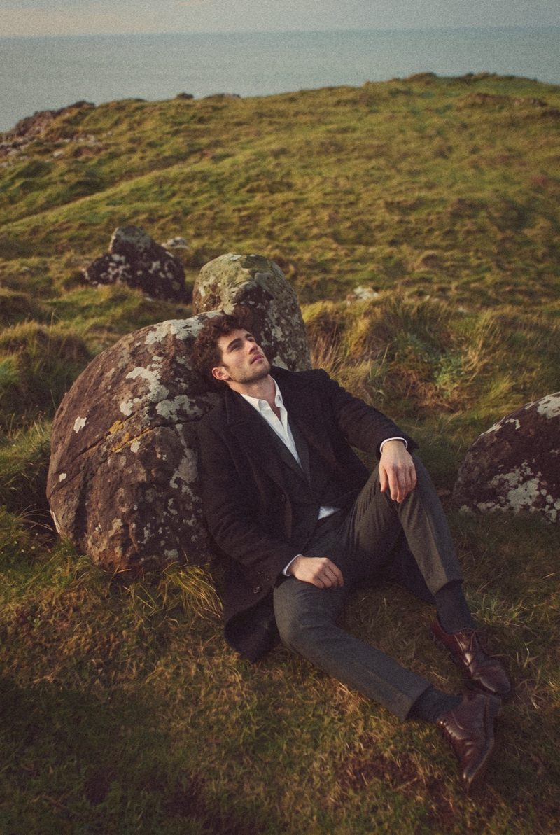 A man in a black overcoat and grey suit leans back against a boulder on a clifftop in low sun.