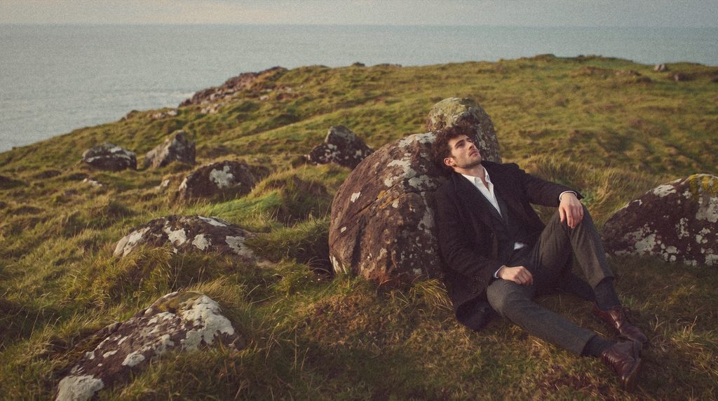 A man in a black overcoat and grey suit leans back against a boulder on a clifftop.