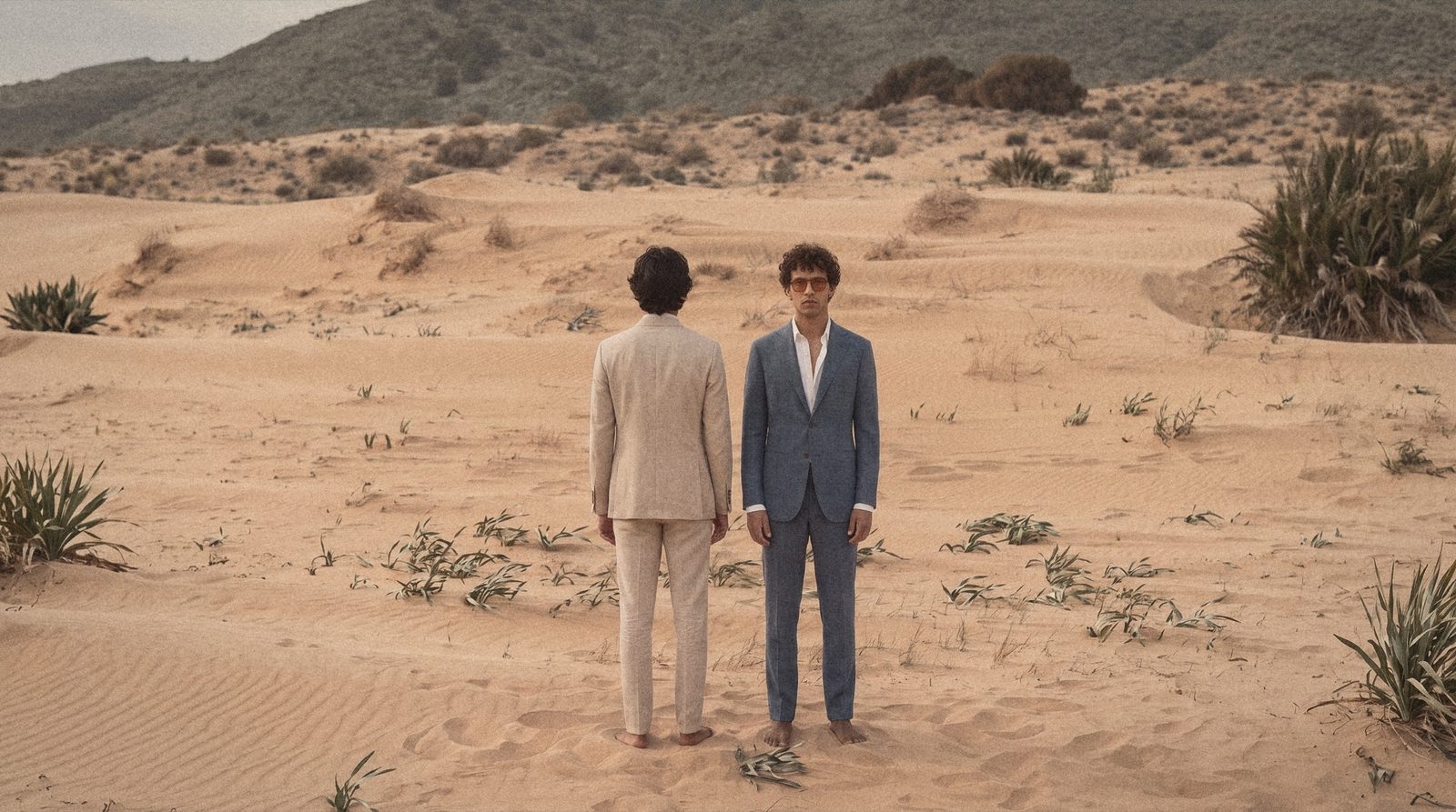 Two men in a sand herringbone suit and a blue check suit stand barefoot in the dunes.