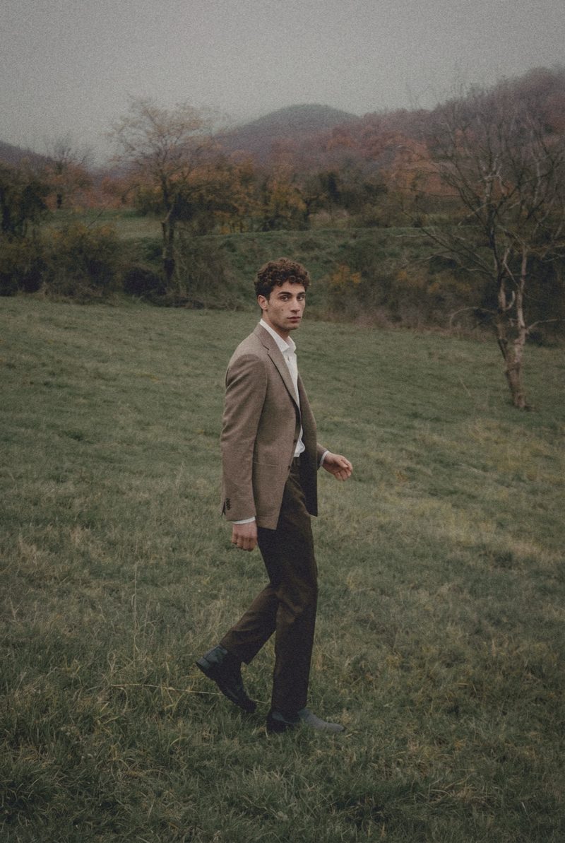 A man in a taupe jacket walks across a foggy meadow and looks back.