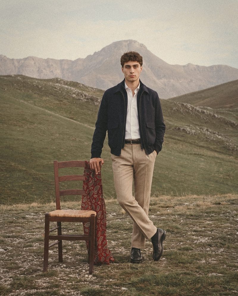 A man in a navy zip jacket and sand trousers stands beside a rush seat chair on a mountain plateau.