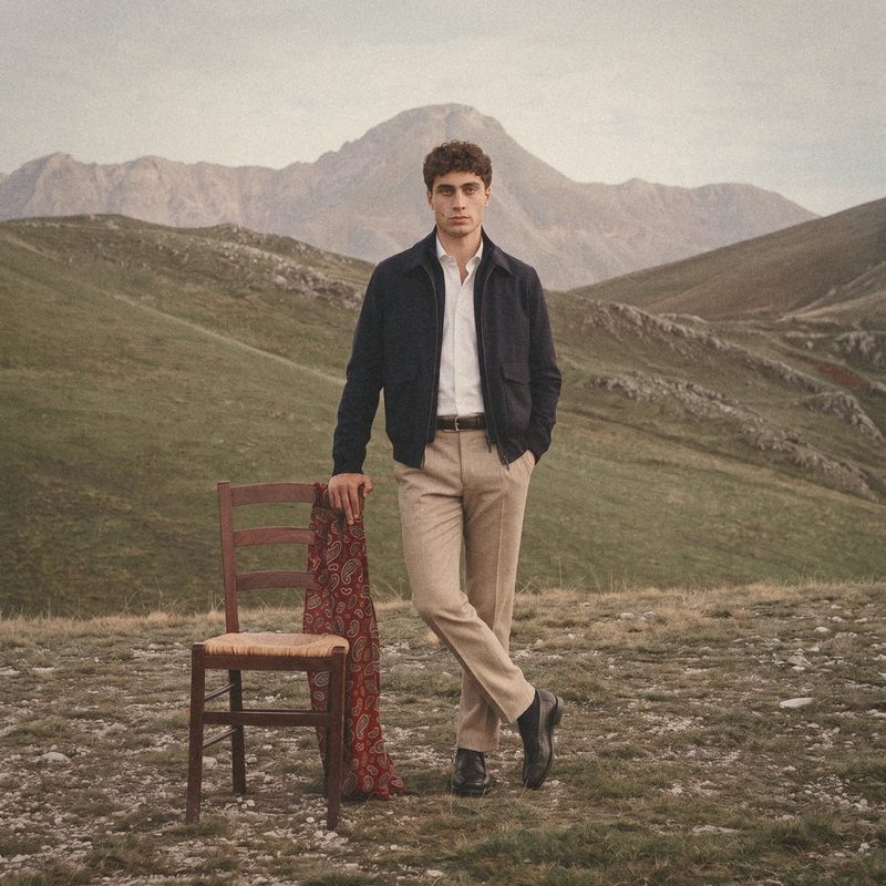 A man in a navy zip jacket and sand trousers beside a rush seat chair on a mountain plateau.