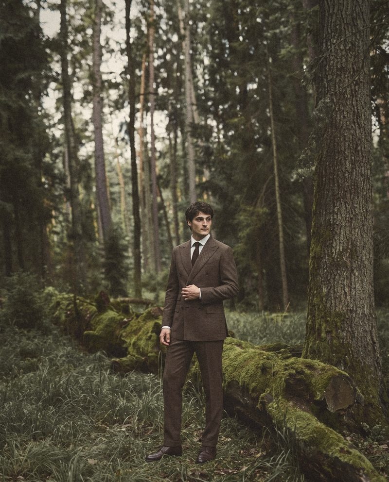A man in a brown houndstooth double breasted suit stands by a mossy fallen trunk.