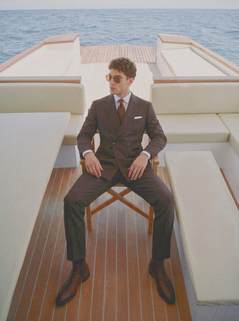 A man in a brown double breasted suit sits on the stern deck of a yacht.