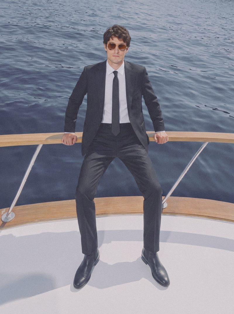 A man in a dark suit and tie sits against the bow rail, the sea behind.