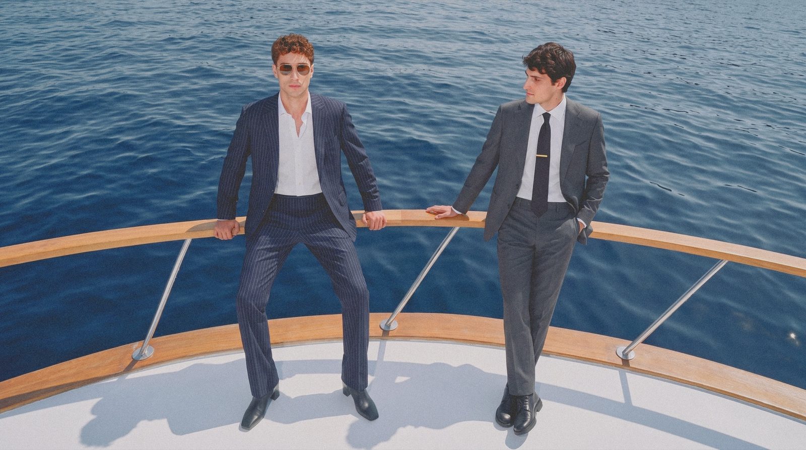 Two men in dark suits lean on the bow rail of a yacht above a blue sea.