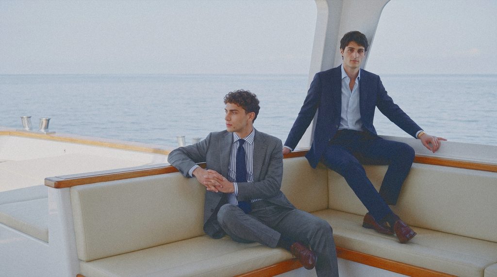 Two men in a grey suit and a navy suit sit on the cream cushions of a yacht cabin.