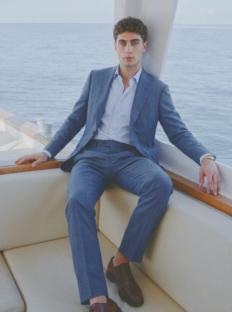 A man in a blue suit and open shirt sits in the cabin corner of a yacht.