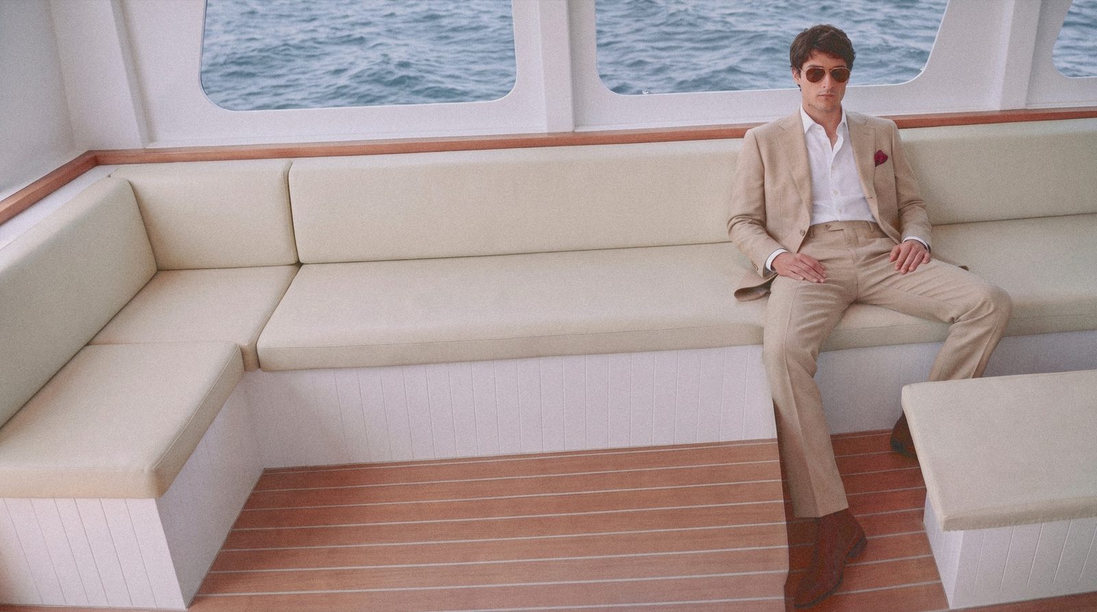 A man in a sand suit sits on the saloon banquette of a yacht, under the windows.